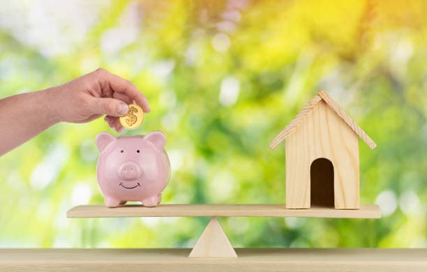 Hand dropping a coin into a piggy bank balanced on a seesaw against a small wooden house